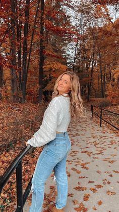 a woman leaning on a rail in front of trees with leaves all over the ground