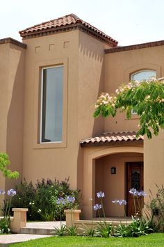 a tan house with purple flowers in the front yard and green grass on the lawn