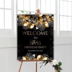 a black and gold birthday party welcome sign on an easel with flowers in the foreground