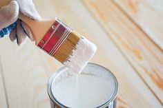 a person holding a paintbrush over a can of white paint on a wooden floor