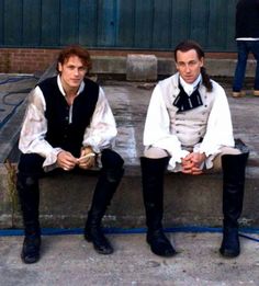 two young men sitting on concrete steps in front of an industrial building, one wearing a vest and tie