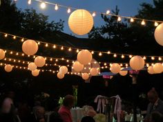 several paper lanterns hanging from the ceiling over tables at an outdoor party with people sitting and standing under them