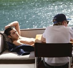 two men sitting at a table next to each other on a boat with water in the background