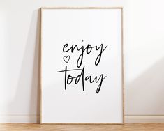 a white framed poster with the words enjoy today on it in black ink next to a wooden floor