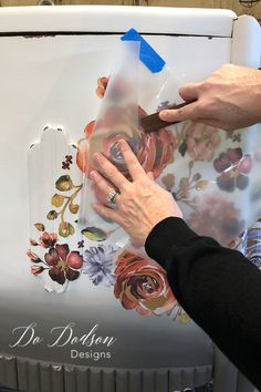 a woman is painting the side of a refrigerator with flowers on it and blue tape