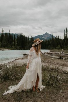 a woman in a white dress and hat standing on the grass near a lake with trees