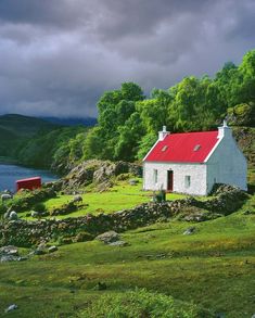 a white house with a red roof sitting on top of a lush green hillside next to a body of water