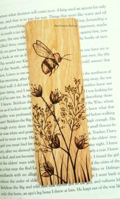 a wooden bookmark with flowers and a bee on it's side, sitting on top of an open book