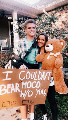 a man and woman standing next to each other holding a sign with a teddy bear