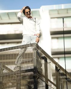 a man standing on top of a metal fence next to a tall building and wearing sunglasses