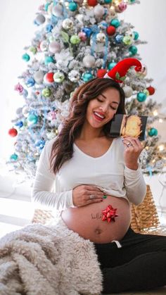 a pregnant woman sitting in front of a christmas tree while holding a piece of bread