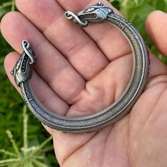 a hand holding a silver bracelet with two dragon heads on it's end, in front of some green plants