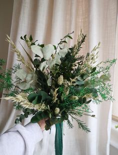 a green vase with white flowers and greenery in front of a curtained window