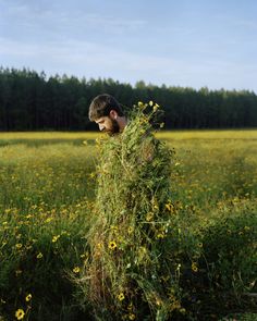 a man standing in a field with flowers on his head and looking down at the ground