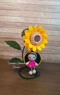 a crocheted doll holding a sunflower on top of a wooden table next to a wall