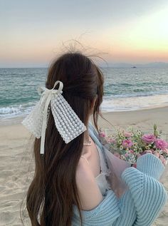 a woman with long brown hair wearing a blue sweater and holding flowers on the beach