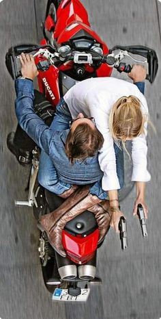 two people are sitting on a motorcycle looking down at the camera man and woman standing next to each other