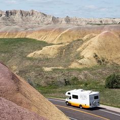 an rv is driving down the road in front of mountains and hills with grass on both sides