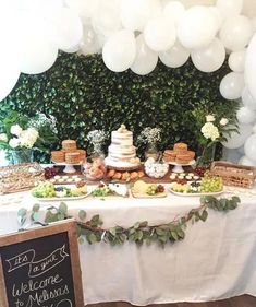 a table topped with lots of desserts and balloons next to a wall covered in greenery