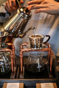 a person pours coffee from a copper kettle into a glass cup on top of a wooden table