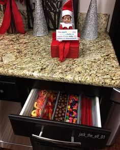 an elf is sitting on top of a counter with candy in the bottom drawer, and another elf has his name sign above it