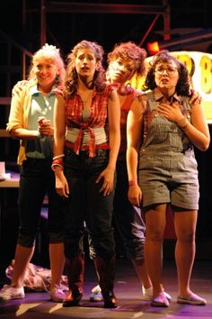 five young women standing on stage with one holding her hand up to her chest and the other looking down