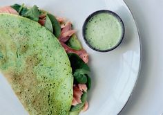 a white plate topped with two tacos covered in meat and veggies next to a cup of green sauce