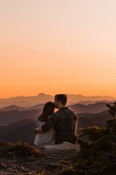 a man and woman sitting on top of a mountain looking out at the mountains in the distance