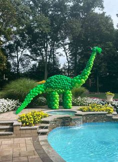 an inflatable dinosaur statue next to a swimming pool with flowers and trees around it