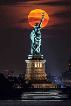 the statue of liberty is lit up at night in front of an orange full moon