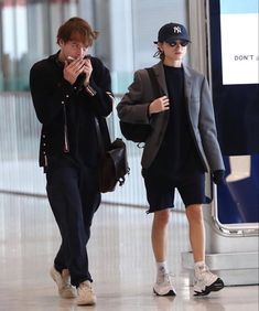 two people are walking through an airport with their cell phones in their hands and one is wearing a baseball cap