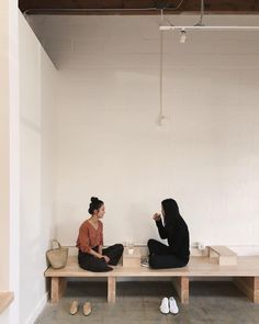 two women sitting on a bench in a room with white walls and flooring, one holding a coffee cup while the other is eating