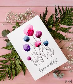 a happy birthday card with flowers and greenery on a pink wooden table next to it