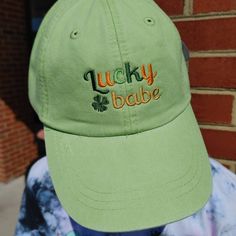 a green lucky babe hat sitting on top of a person's head in front of a brick wall