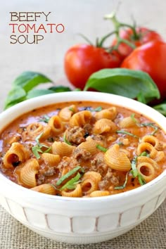 a white bowl filled with pasta and meat soup on top of a table next to tomatoes