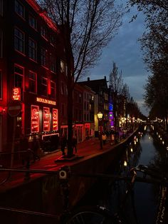 people walking down the street at night with red lights on buildings and water in front of them