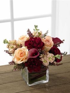 a square vase filled with flowers on top of a wooden table next to a window