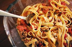 a glass bowl filled with pasta and meat on top of a wooden table next to a fork