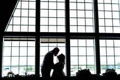 a bride and groom standing in front of large windows at their wedding reception, silhouetted against the backdrop of an airplane landing strip