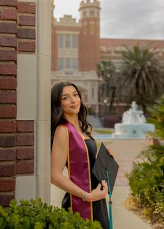 a woman in a graduation gown standing next to a brick building holding a black folder