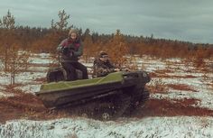 two people riding on the back of an army vehicle in snow covered field next to trees