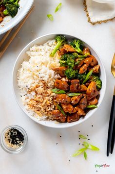 a white bowl filled with rice, broccoli and chicken next to chopsticks