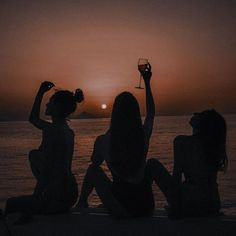 two women sitting on the beach at sunset holding wine glasses in front of their faces