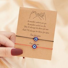 a hand holding a card with two evil eyes on it and an orange string bracelet that says, no matter where we go