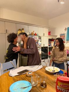 three people standing around a kitchen table with plates and cups on it, one man is kissing the other