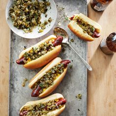 three hot dogs on buns with toppings next to a bowl of mustard and two spoons