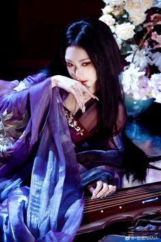 a woman with long black hair sitting next to a purple guitar and flowers in the background