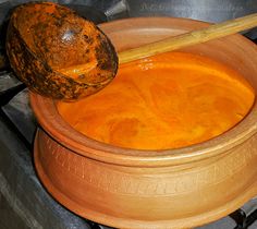 a wooden spoon in a clay pot filled with orange colored liquid next to a coconut