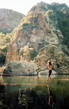 a boy is walking across the water in front of some mountains and rocks, while holding onto a rope