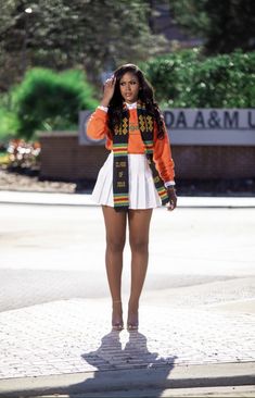 a woman in an orange jacket and white dress is standing on the sidewalk with her hands behind her head